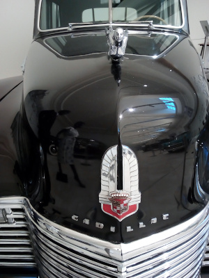 [The car is black with a silver grill. Just above the grill is the word CADILLAC in individual sliver letters. The mostly red and white Cadillac crest and badge emblem is just above that. On the top of the hood is a silver flying lady hood goddess.]